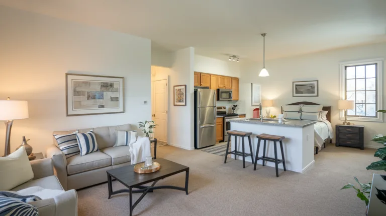 A photo of a large, well-lit above garage apartment with a living room, kitchen, and bedroom. The living room has a sofa, a coffee table, and a lamp. The kitchen has a refrigerator, a stove, and a sink. The bedroom has a bed, a nightstand, and a lamp. The walls are painted white, and the floors are covered with beige carpeting. There is a window in the bedroom.